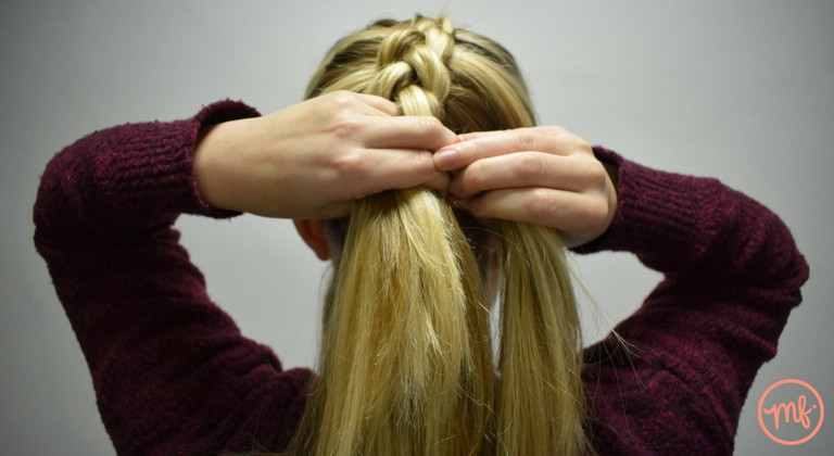 Blonde haired girl wearing a red jumper doing a dutch braid on herself