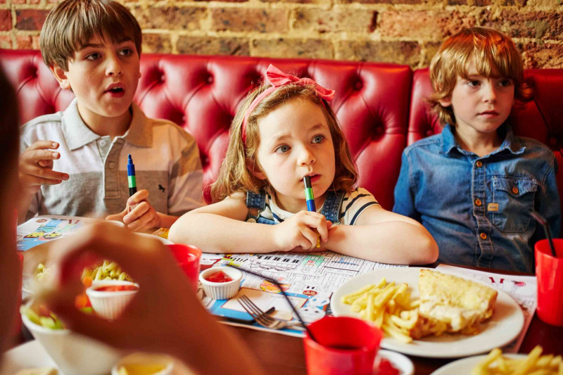 3 kids sat on a red cushioned bench at Cafe Rouge, crayons in hand
