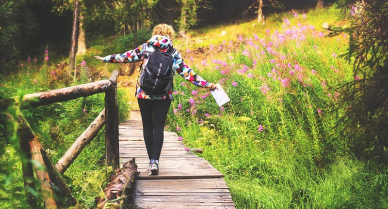 Back of woman walking across a wooden pathway, holding a map with a backpack on. Walking through a green woodland with flowers and trees all around