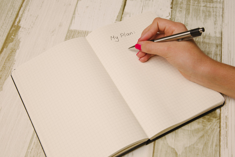 White female hand with pink nail varnish holding a black pen about to write in a notepad, on a page with the title 'My Plan'