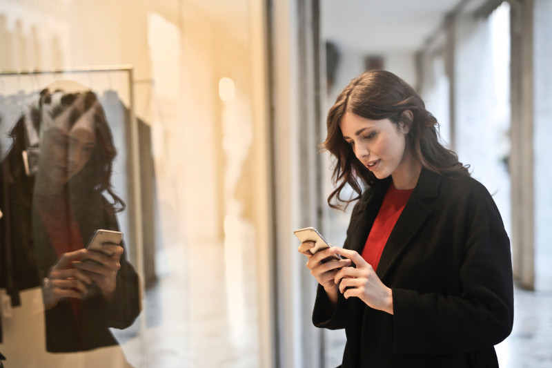 Brunette woman wearing red jumper and black coat looking at iPhone with reflection in mirror