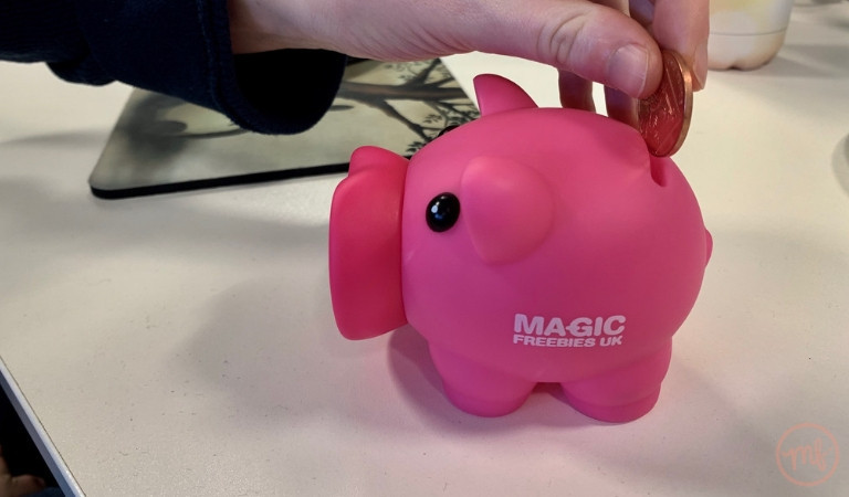 Hand putting 2 pence in pink Magic Freebies piggy bank